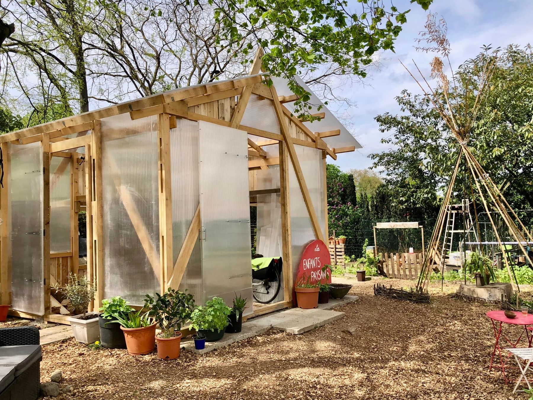 L’Ambassade des fruits et légumes : une serre en bois construite en chantier participatif, entourée de plants en pots et d’un tipi de bambou